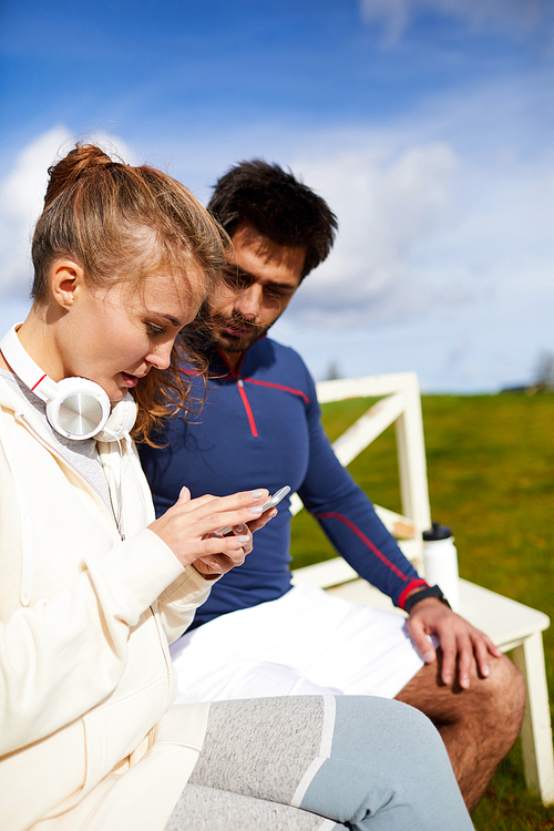 Young active woman scrolling in her smartphone while sitting next to her boyfriend outdoors