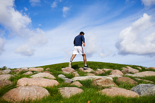 Young man in sportswear walking down big stones on green field as part of his morning workout