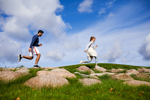 Active guy and girl running down large stones on green field against cloudy sky