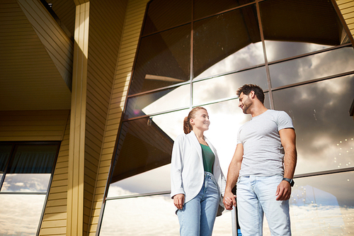 Young contemporary couple in casualwear holding by hands by large window of modern architecture outside