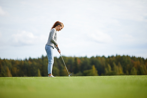 Young active woman in casualwear standing on golf field and getting ready to hit ball with club