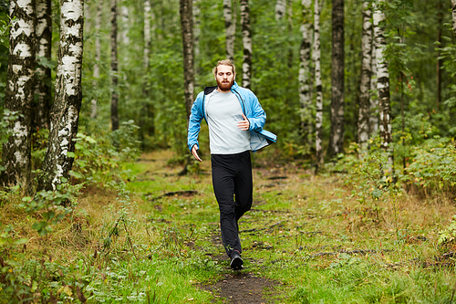 Young active man running down forest path surrounded by green grass and trees on summer weekend