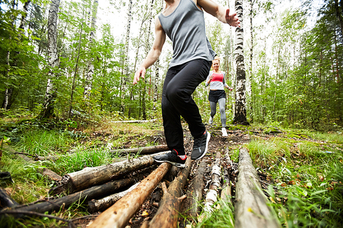 Young active man and woman in sportswear moving down logs on forest path while training in the morning