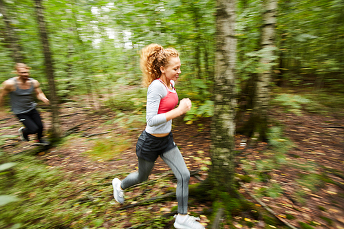 Young active female and her boyfriend running in birch tree forest around blurry green foliage in the morning