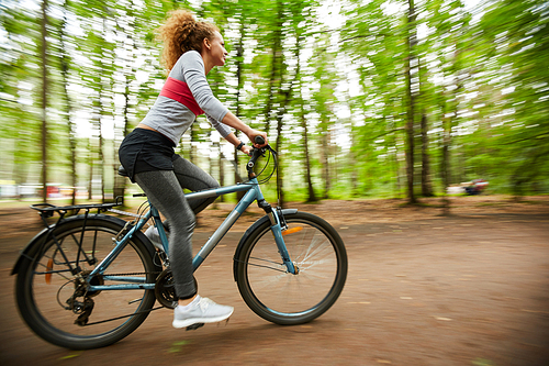 Young sportswoman riding bicycle along road in the forest with blurry trees on both sides