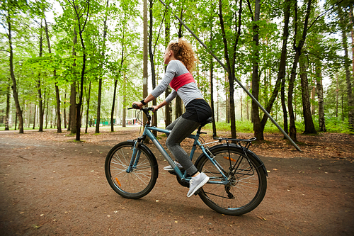 Active girl in sportswear sitting on bicycle while moving down forest road on summer day or weekend