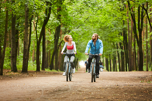 Young man and woman in activewear talking while cycling on road in park on summer weekend