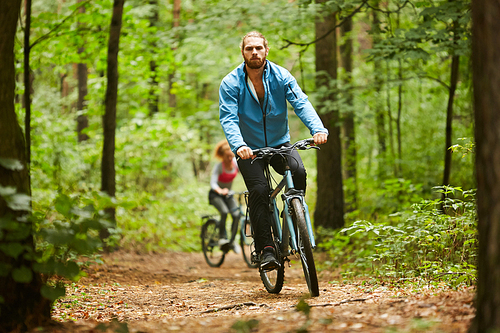 Active guy in sportswear cycling along forest path on summer day with his girlfriend moving after him