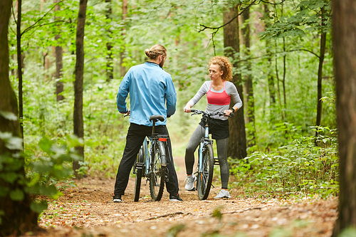 Active girl and guy with bicycles standing on forest path and discussing what way to choose for ride