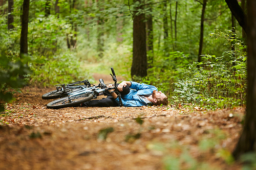 Active young man holding by his hurt or broken leg while lying on forest path by his bicycle