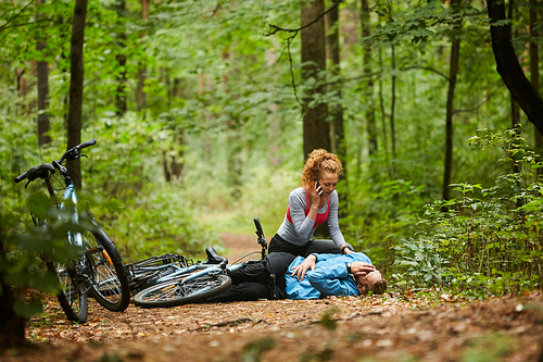 Young woman calling ambulance in the forest while leaning over her boyfriend lying on path with his leg broken