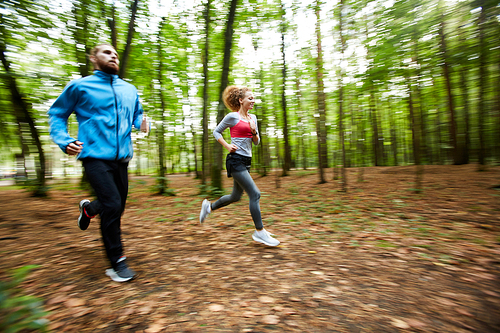 Young man in activewear and his girlfriend running together in the forest during morning chill