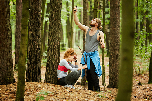 Young man with smartphone catching signal to call for help while sad woman with hit leg sitting by tree