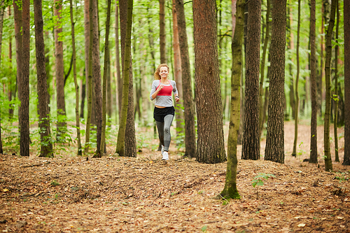 Young sportswoman running down forest path among trees while training on summer morning