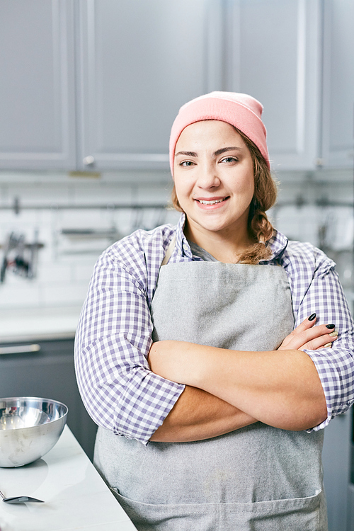 Portrait of young Caucasian female cook in apron standing in kitchen with her hands crossed and smiling at camera