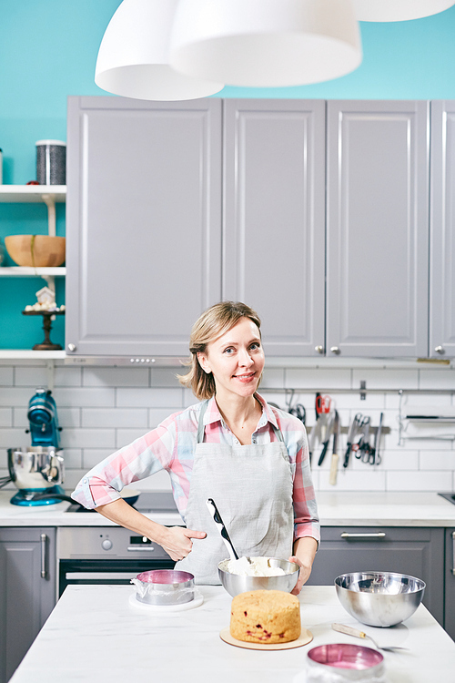 Young attractive Caucasian woman in apron standing in kitchen with freshly baked pastry and smiling at camera