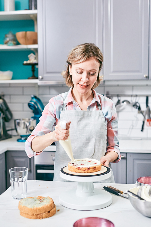 Attractive Caucasian woman using pastry bag while decorating appetizing cake with whipped cream in kitchen