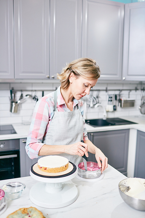 Young blond-haired Caucasian woman in apron making delicious cake in kitchen