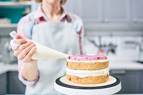 Unrecognizable female pastry cook squeezing strawberry cream on appetizing layer cake in kitchen