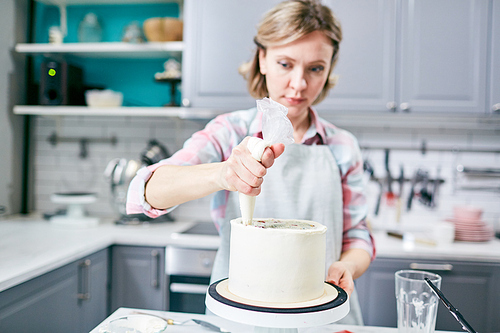 Defocused young Caucasian woman using pastry bag while covering appetizing cake with frosting in kitchen