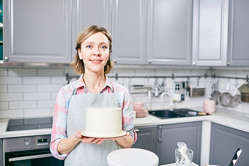 Portrait of attractive Caucasian woman holding delicious frosted cake in kitchen and smiling at camera cheerfully