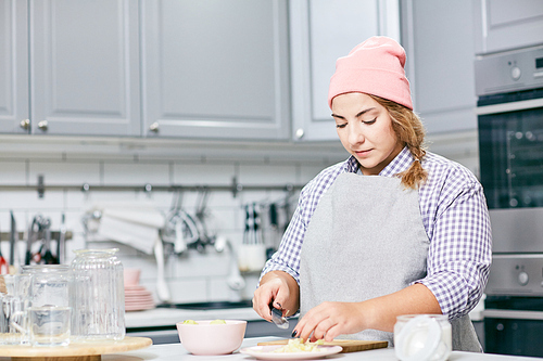 Young concentrated Caucasian woman in apron and hat cutting pear in modern kitchen