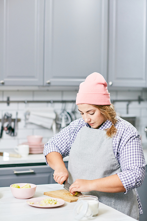 Stylish plump Caucasian woman wearing apron and hat cutting pear with chef knife in modern kitchen