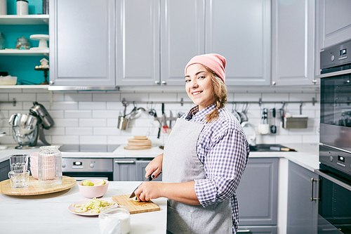 Young pretty female cook in apron and hat making fruit salad in modern kitchen and smiling at camera happily