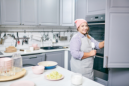 Portrait of pretty Caucasian woman in apron and hat standing by open fridge in modern kitchen and smiling at camera