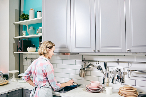 Rear view of blond-haired woman standing by stove in modern kitchen and cooking dish in frying pan