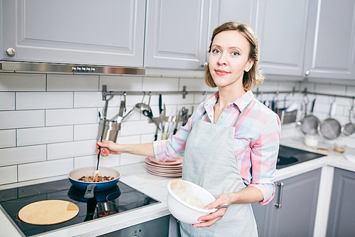 Portrait of attractive blond woman standing near electric stove, stirring food in frying pan and smiling at camera