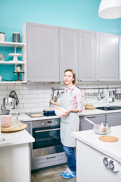 Full-length portrait of Caucasian woman standing by electric range, preparing meal in frying pan and 