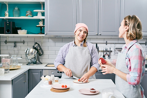 Young stylish Caucasian female chef cutting strawberries in kitchen, talking to her colleague and smiling