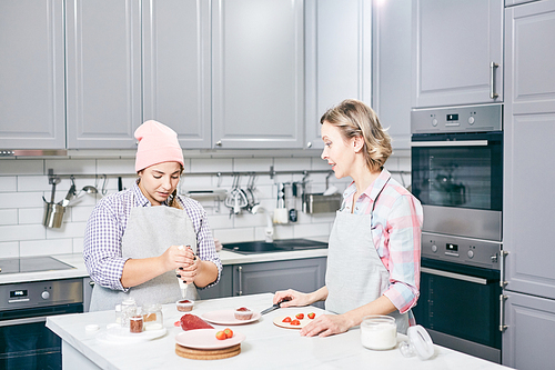 Young Caucasian women in aprons preparing appetizing strawberry cupcakes  in modern kitchen