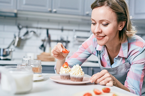 Attractive Caucasian female pastry cook decorating delicious cupcakes with fresh strawberries and smiling happily