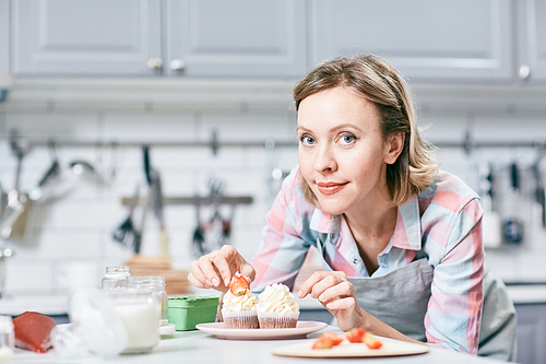 Portrait of attractive Caucasian woman decorating appetizing cupcakes with strawberries and smiling at camera