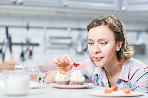 Portrait of blond Caucasian female cook decorating tasty cream cupcakes with fresh berries in kitchen