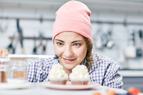 Young female pastry cook in pink hat decorating cream cupcakes with blueberries on blurred background