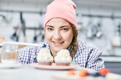 Portrait of young woman in pink hat smiling at camera while decorating appetizing cream cupcakes on blurred background