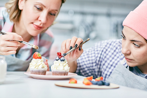 Young concentrated Caucasian women decorating cream cupcakes with fresh strawberries, blueberries and currants