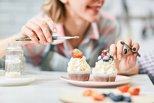 Unrecognizable female pastry cooks decorating delicious cream cupcakes with fresh berries and frosting, close-up view