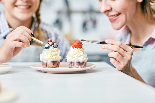 Close-up view of Caucasian women smiling cheerfully while decorating appetizing cream cupcakes with berries and frosting