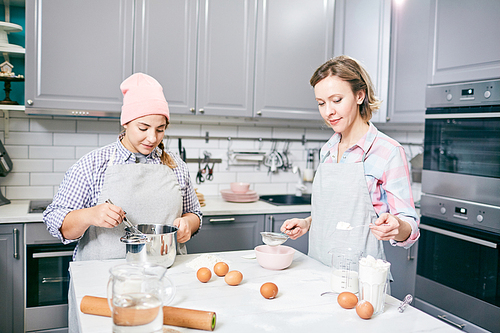 Young Caucasian women whisking eggs and sifting flour through sieve while cooking dessert in modern kitchen