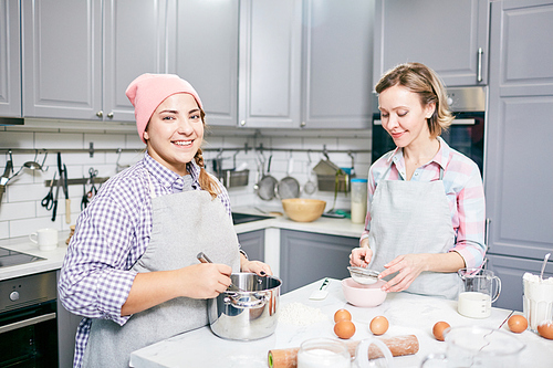 Portrait of young pretty woman standing in kitchen, whisking eggs for cake and smiling at camera while her female friend sifting flour