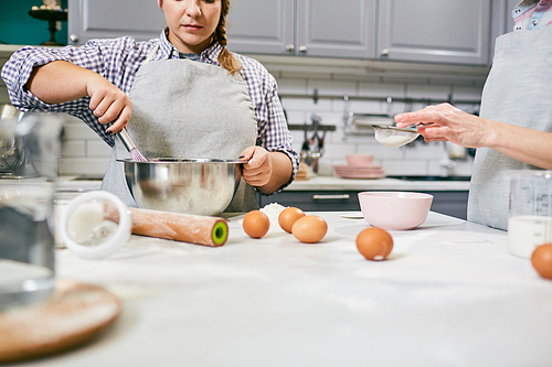 Low angle view of young female cook and her unrecognizable colleague whisking eggs and sifting flour through sieve in kitchen