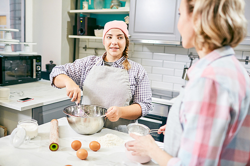 Young Caucasian female cook in pink hat whisking eggs for pastry and listening to her colleague