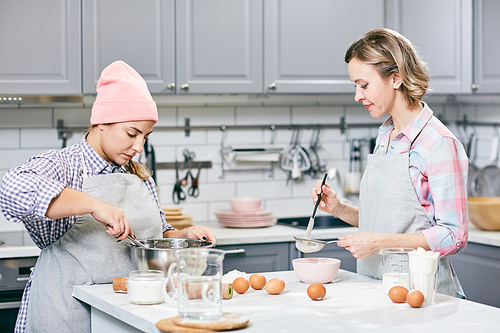 Professional female confectioners whisking eggs and sifting flour through sieve while making cake in modern kitchen