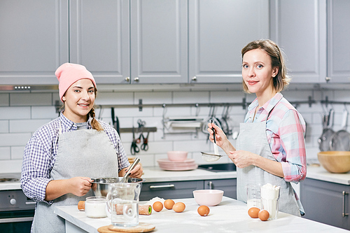 Portrait of young attractive female cooks smiling at camera while whisking eggs and sifting flour for cake in kitchen