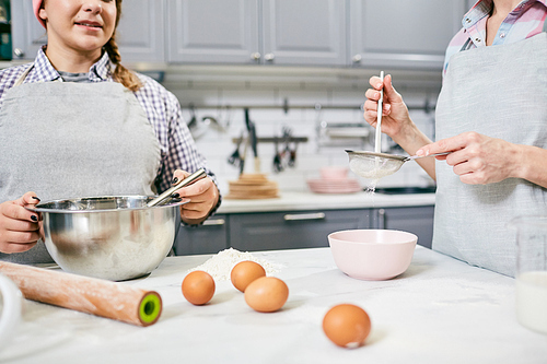 Young female pastry cook and her unrecognizable colleague whisking eggs and sifting flour through sieve in kitchen