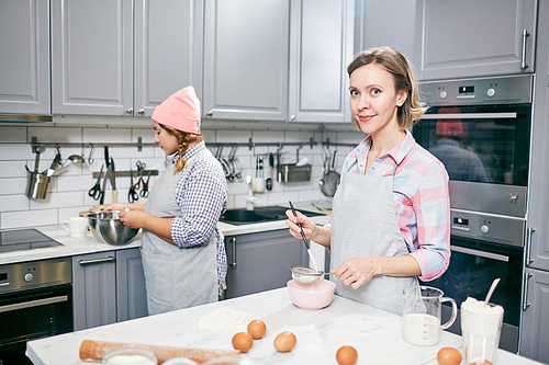 Attractive Caucasian female cook sifting flour through sieve and smiling at camera while her colleague whisking eggs